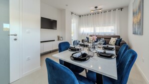 A modern dining area with a white table set for four, blue upholstered chairs, and a large window with sheer curtains. - Two-bedroom apartment with a terrace (Novalja)