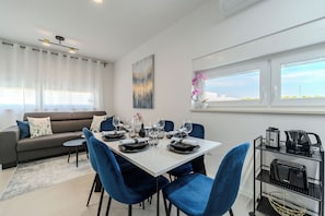Un comedor moderno con una mesa blanca para cuatro, sillas azules, un sofá gris con cojines decorativos, y una cocina con electrodomésticos sobre un carrito metálico.
