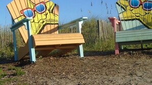 Dos sillas Adirondack coloridas con caras caricaturescas y gafas de sol, ubicadas en una zona arenosa con césped y una cerca al fondo.