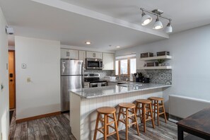 A modern kitchen with a central island, bar stools, stainless steel appliances, and a backsplash with patterned tiles. - NEW Ski in/out condo with Mt views, hot tub, porch, gym & wifi! (Dover)
