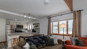 Une cuisine moderne équipée d’appareils électroménagers en acier inoxydable, un coin repas avec une table en bois et des tabourets de bar, et un salon confortable avec un canapé et une lampe.