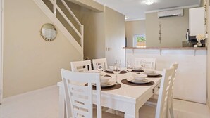 A dining area with a white table set for four, surrounded by white chairs. A staircase is visible to the left, and an air conditioning unit is mounted on the wall above the counter. - Luxurious 2-Bedroom Condo near Eagle Beach - Noord (Noord)