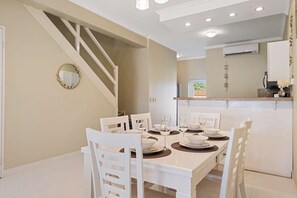 A dining area with a white table set for four, surrounded by white chairs. A staircase is visible to the left, and an air conditioning unit is mounted on the wall above the counter. - Luxurious 2-Bedroom Condo near Eagle Beach - Noord (Noord)