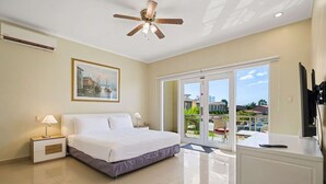 A hotel room with a bed, a desk, a ceiling fan, a painting, and a view of the outside. - Luxurious 2-Bedroom Condo near Eagle Beach - Noord (Noord)