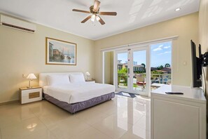 A hotel room with a bed, a desk, a ceiling fan, a painting, and a view of the outside. - Luxurious 2-Bedroom Condo near Eagle Beach - Noord (Noord)