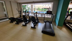 Une salle de sport équipée de matériel d’exercice, d’un tapis de course, et offrant une vue sur une piscine et les bâtiments environnants.