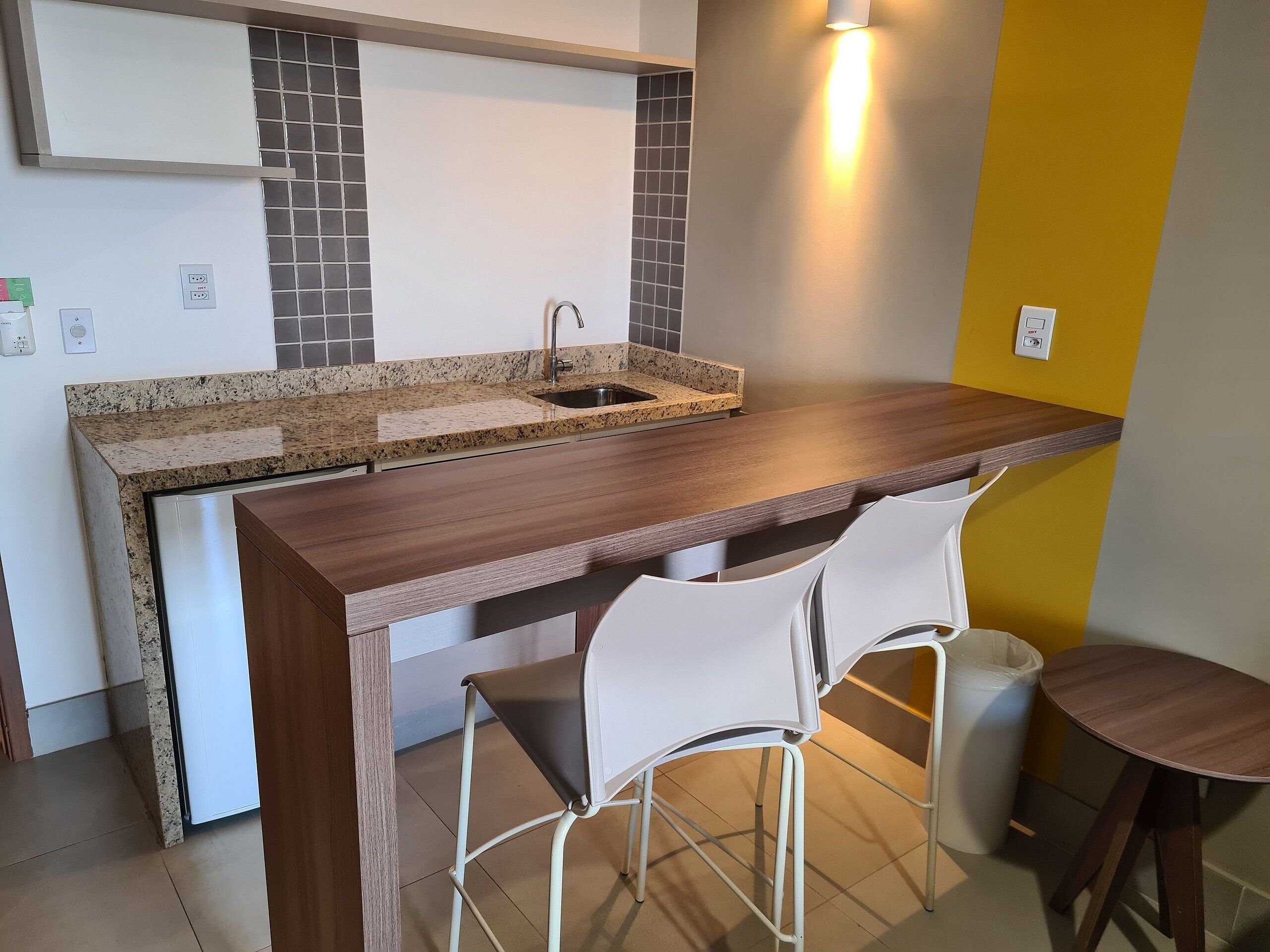Een keuken met een granieten aanrecht, een roestvrijstalen spoelbak en een gele accentmuur. Er staan twee witte stoelen en een kleine tafel.