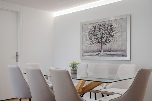 A modern dining area with a glass table, white chairs, and a painting of a tree. - Athens Golden Light Premium Residence (Athens)