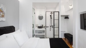 A modern bedroom with a bed, a black bench, a walk-in closet, and a bathroom with a shower and sink. - Athens Golden Light Premium Residence (Athens)