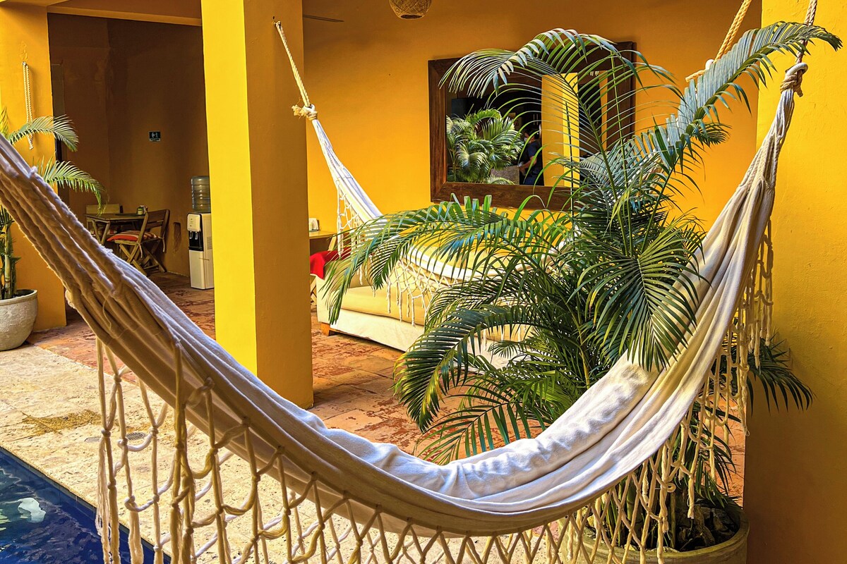 A hammock tied between two pillars in a yellow-walled courtyard with a ceiling fan, a water dispenser, and a potted plant.