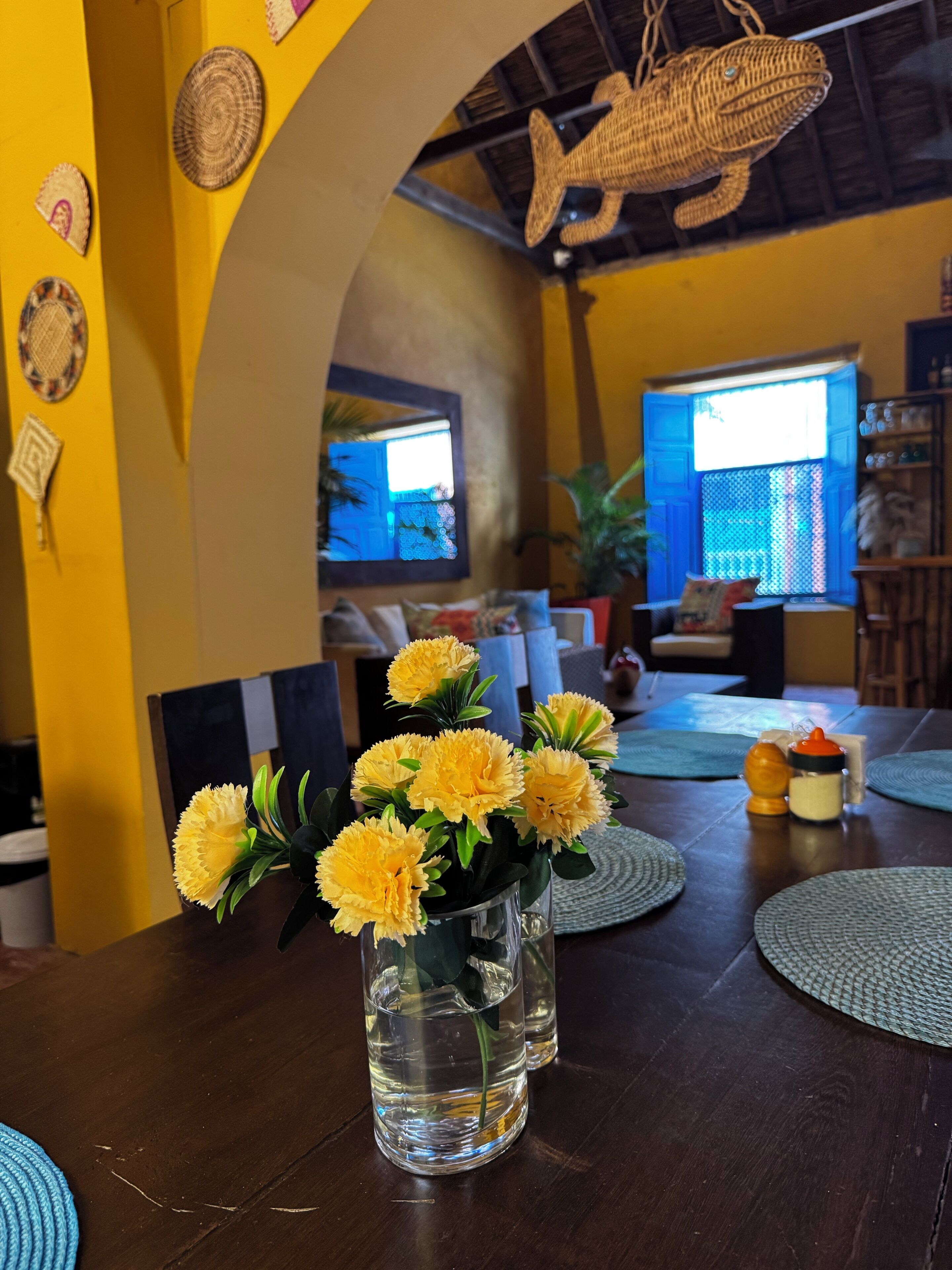 Une table à manger ornée d’un vase de fleurs jaunes, une table en bois et un set de table bleu à motifs.