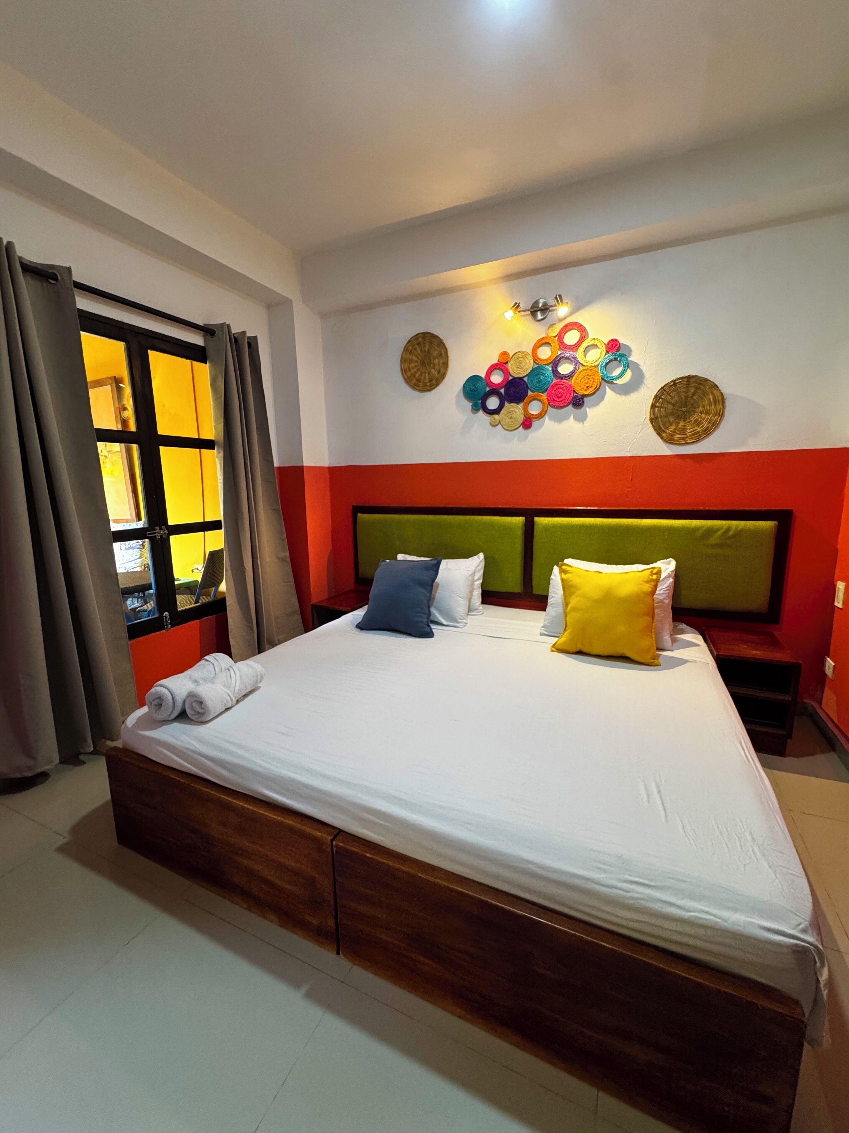 Une chambre d’hôtel avec un grand lit, une fenêtre avec des rideaux, un mur orné d’un motif circulaire coloré et une table de chevet avec une lampe.
