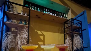 A bar area with three margaritas on a wooden bar counter, shelves with bottles and glasses, and decorative feathers. - CASA ENCANTADA  (Santa Marta)