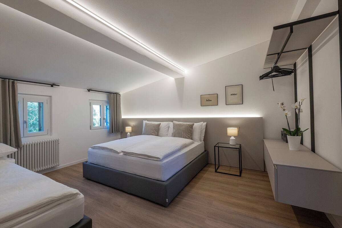 Quarto de hotel moderno com uma cama grande, duas mesas de cabeceira, uma mesinha lateral, um radiador e uma janela com cortinas.
