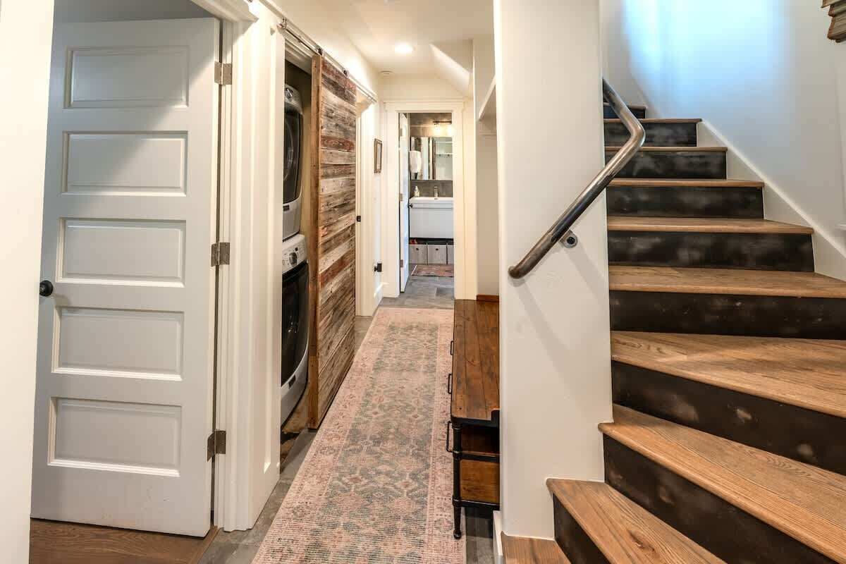 A hallway with a wooden staircase, a patterned rug, and a kitchen area in the distance.