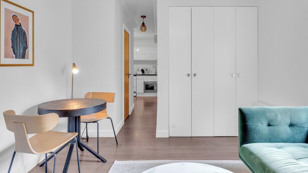 A modern living room with a round dining table, two chairs, a teal sofa, and a kitchen in the background. - 1-Bedroom City Sophistication: Unwind in Style! (London)
