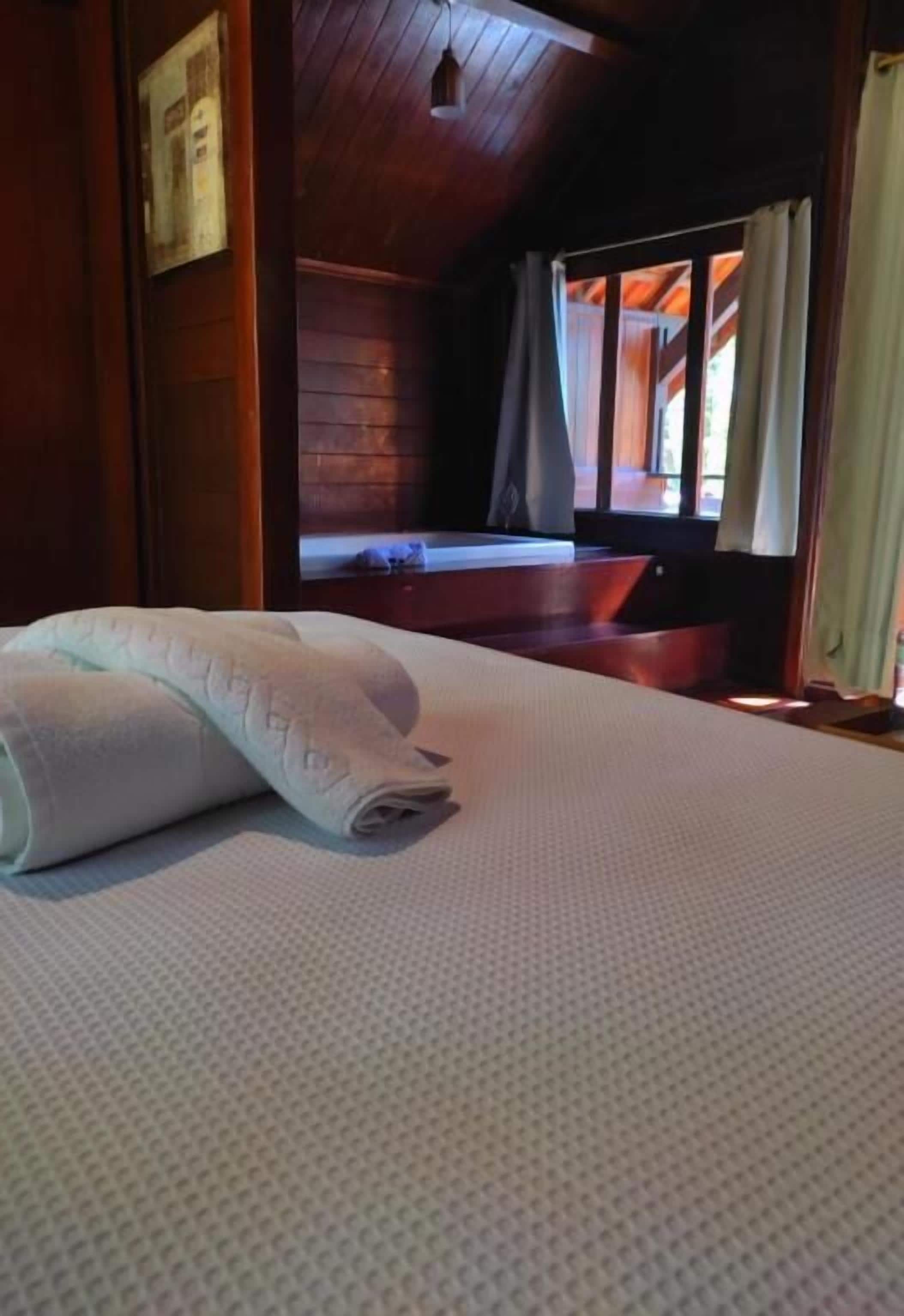 A neatly made bed with a rolled white towel, wooden walls, a window with curtains, and a wooden ceiling.