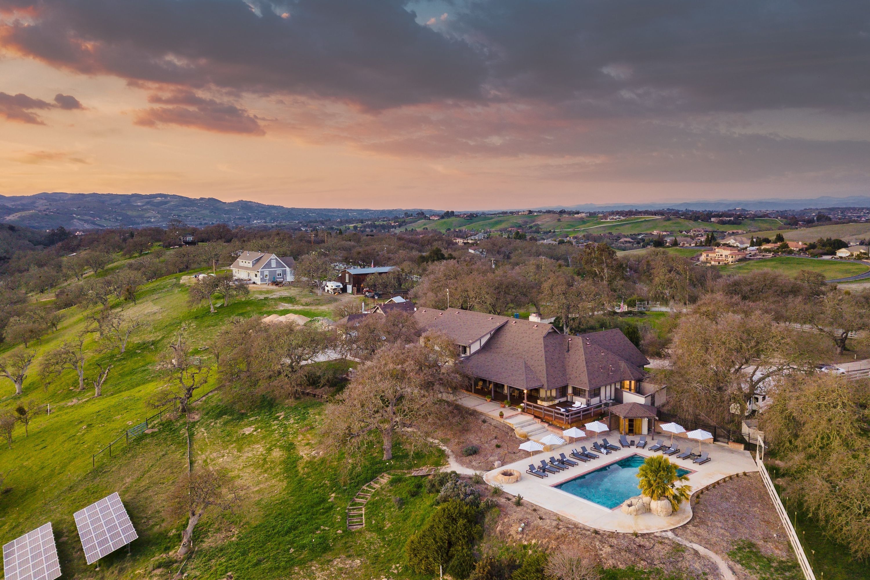 A large house with a swimming pool and solar panels, surrounded by green hills and trees.