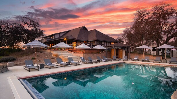A swimming pool with lounge chairs and umbrellas, surrounded by a building with a sloped roof and a view of a sunset. - Cathedral by Avantstay Charming Secluded Estate w Sauna Pool Table Views of the Rolling Hills (Templeton)