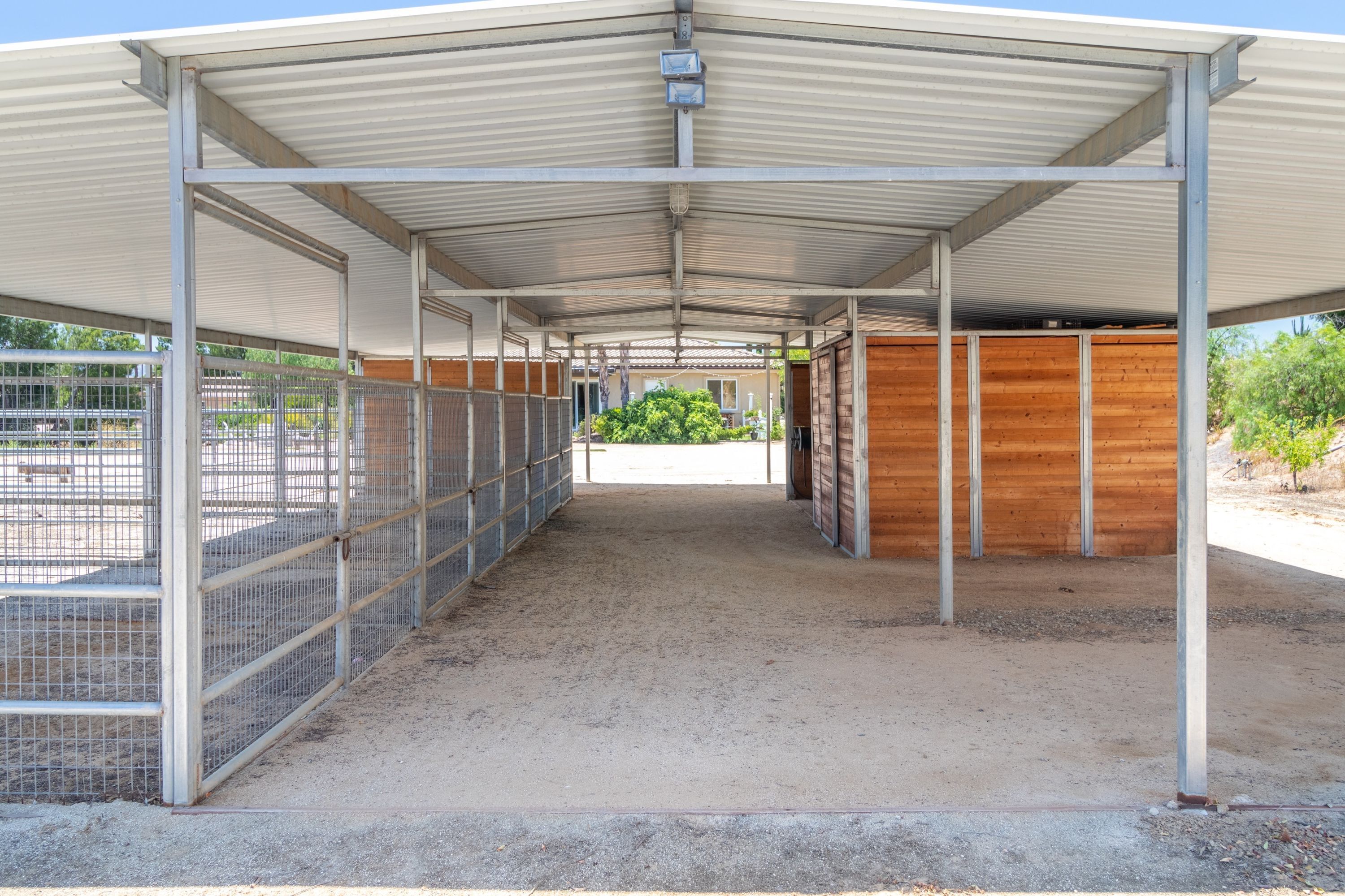 A covered stable with metal fencing, wooden stalls, and a dirt floor.