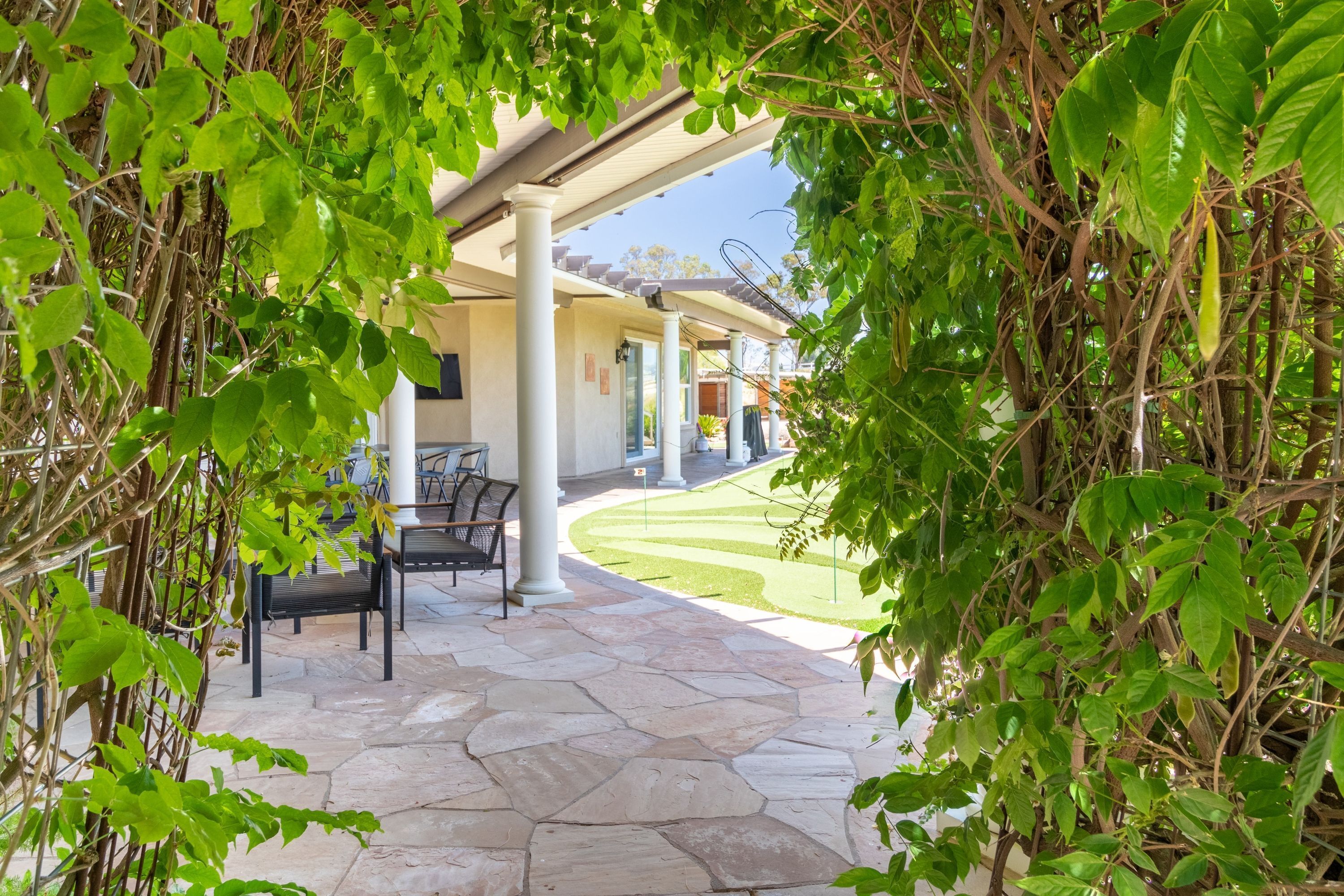 Un patio cubierto con suelo de piedra, mobiliario exterior y abundante vegetación.
