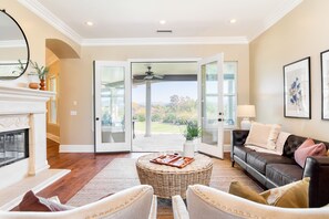 A spacious living room with a fireplace, a comfortable seating area, and a view of the outdoors through glass doors. - Bear Bridge by Avantstay Hilltop Haven in Temecula w Views Putting Green Hot Tub (Temecula)