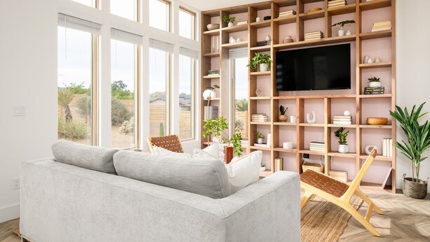 A modern living room with a large window, a grey sofa, a wooden bookshelf, and a television. - Chicory by Avantstay Modern Desert Retreat w Hot Tub (Joshua Tree)