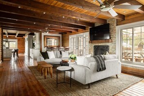 A spacious living room with a fireplace, wooden beams, and a large window overlooking a patio.