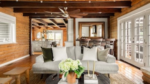 A log cabin interior with a sofa, a coffee table with a vase of flowers, a ceiling fan, and a dining area in the background.