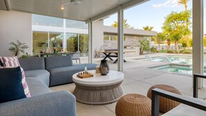 Un soggiorno moderno con un divano ad angolo grigio, un tavolino rotondo, pouf in vimini e vista su una piscina e zona pranzo all'aperto.