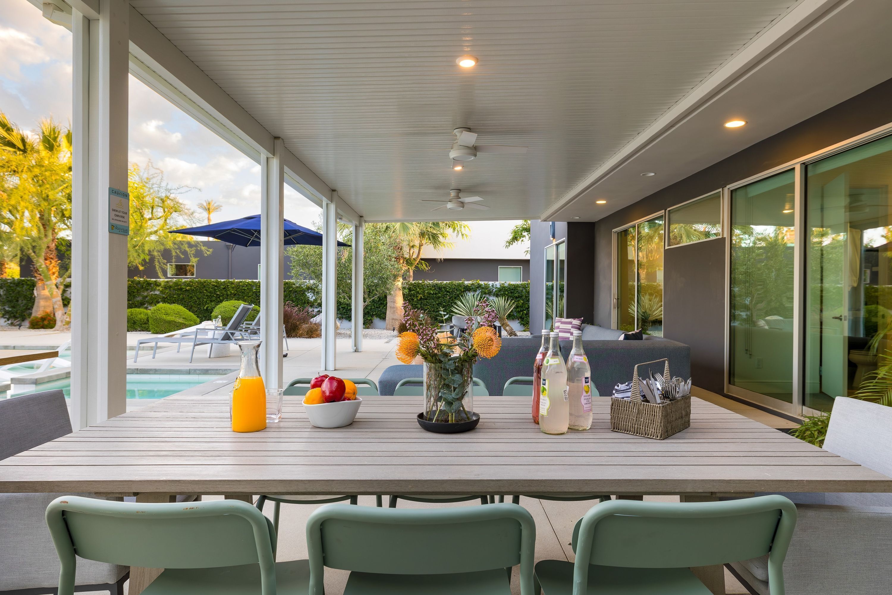 A modern outdoor dining area with a pool view, a table set with fruits, drinks, and a vase of flowers.