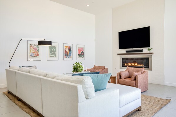 A modern living room with a white sofa, a pink armchair, and a flat-screen TV mounted above a fireplace.