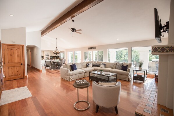 A spacious living room with a large sectional sofa, a coffee table, and a fireplace. There is a dining area in the background with a chandelier. - De Luz by Avantstay 5 Acre Estate w Lake Pool Hot Tub (Temecula)