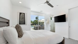 A modern bedroom with a large bed, a ceiling fan, a flat-screen TV, and a view of a pool and outdoor area. - Polo Villa 7 by Avantstay Features Entertainer s Backyard Game Room 260316 5 Bedrooms (La Quinta)