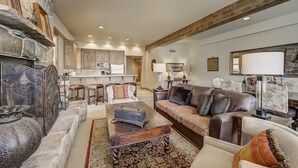 A spacious living room with a fireplace, a sofa, and a coffee table. There is a kitchen in the background with wooden cabinets and a countertop.