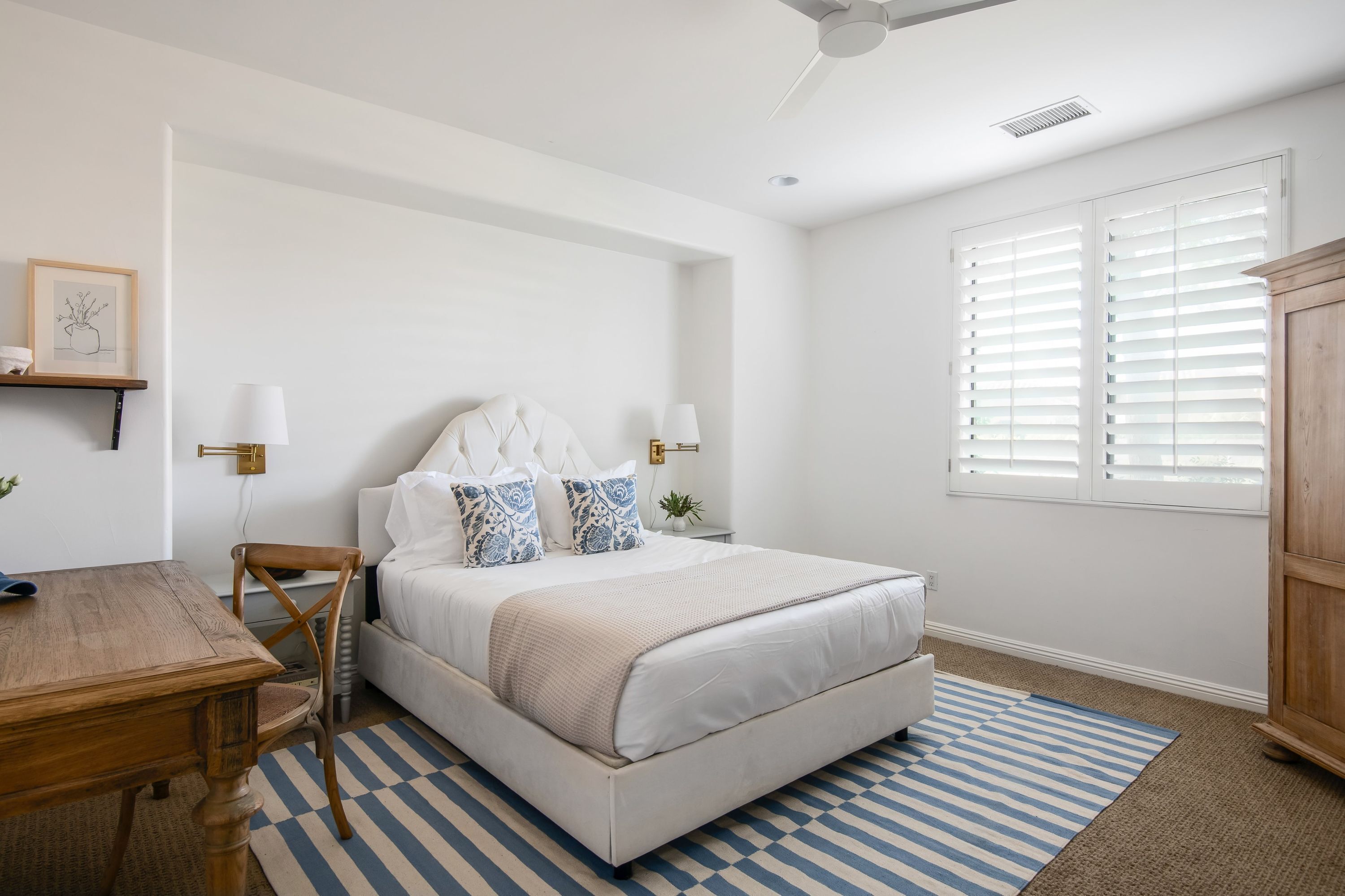 Une chambre à coucher avec un lit, une table en bois, une chaise, un tapis à rayures bleues et blanches, et une fenêtre avec des stores.