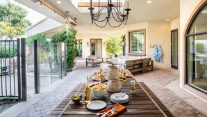 An outdoor dining area with a wooden table set for a meal, surrounded by a patio with a view of greenery and a building. - Villa Verbena by Avantstay Expansive Estate w B-ball Court Pool Spa Entertainer s Yard (Indio)