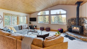 A cozy living room with a stone fireplace, wooden ceiling, and large windows offering a view of trees. - Golden Summit by Avantstay Stunning Secluded Cabin w Access to Tahoe Donner (Truckee)