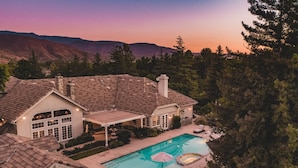 Una casa grande con piscina rodeada de árboles y montañas al fondo.