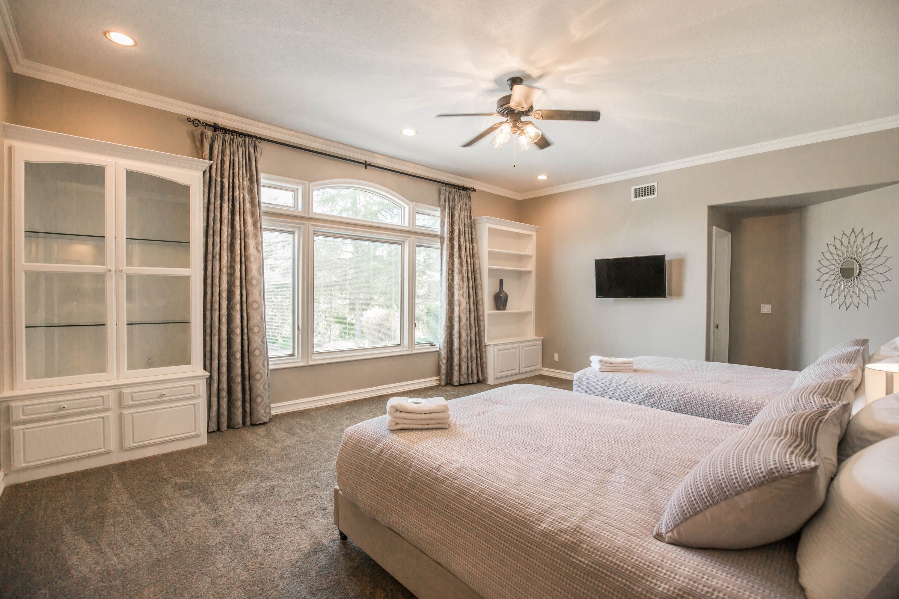 A bedroom with two beds, a ceiling fan, a TV, and a large window with curtains.