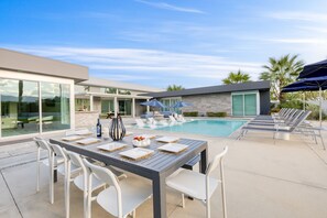 A modern outdoor dining area with a pool, white chairs, and a table set with plates and a bottle.