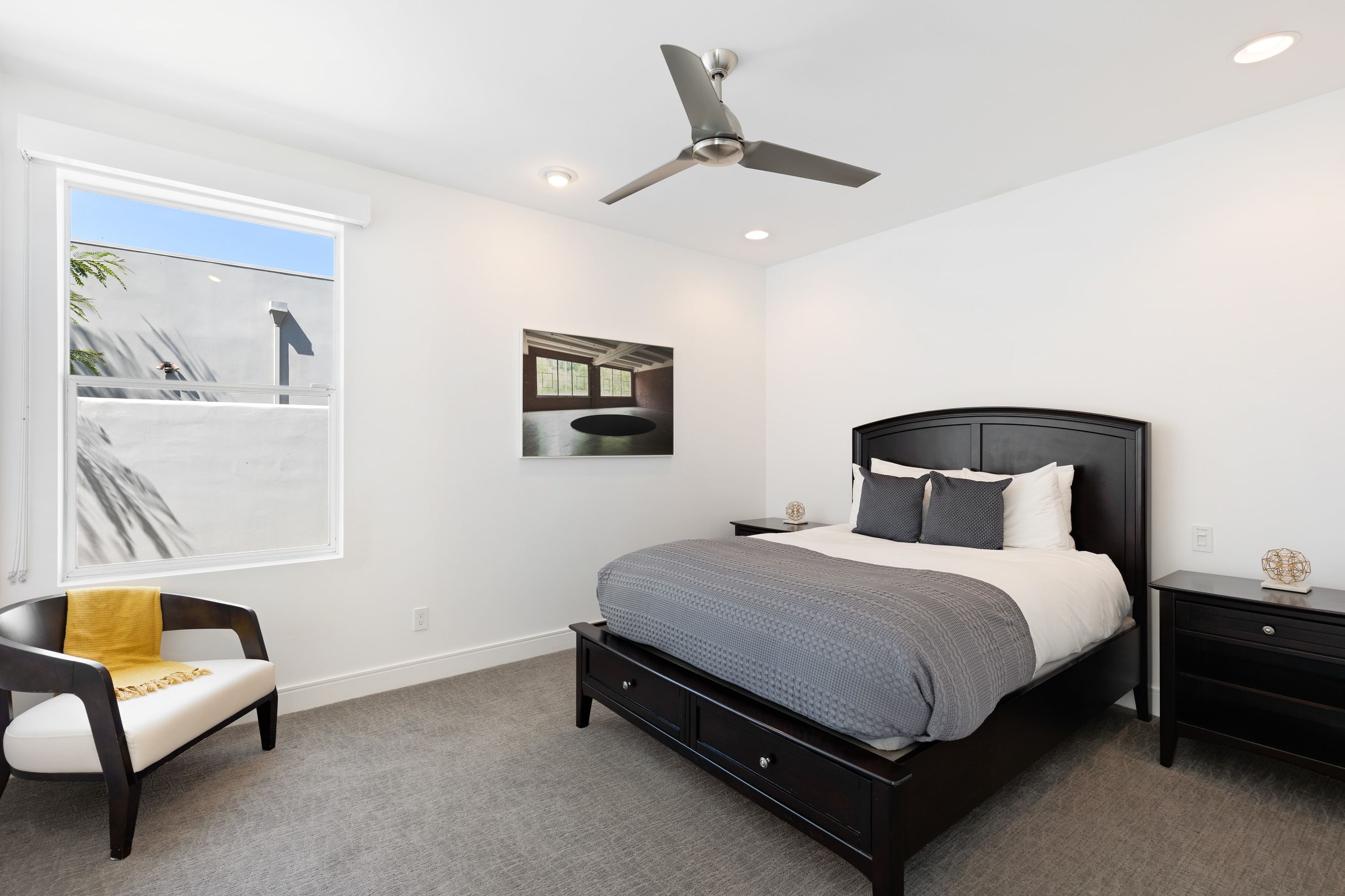 A bedroom with a bed, a chair, a nightstand, a ceiling fan, and a window with a view of palm trees.
