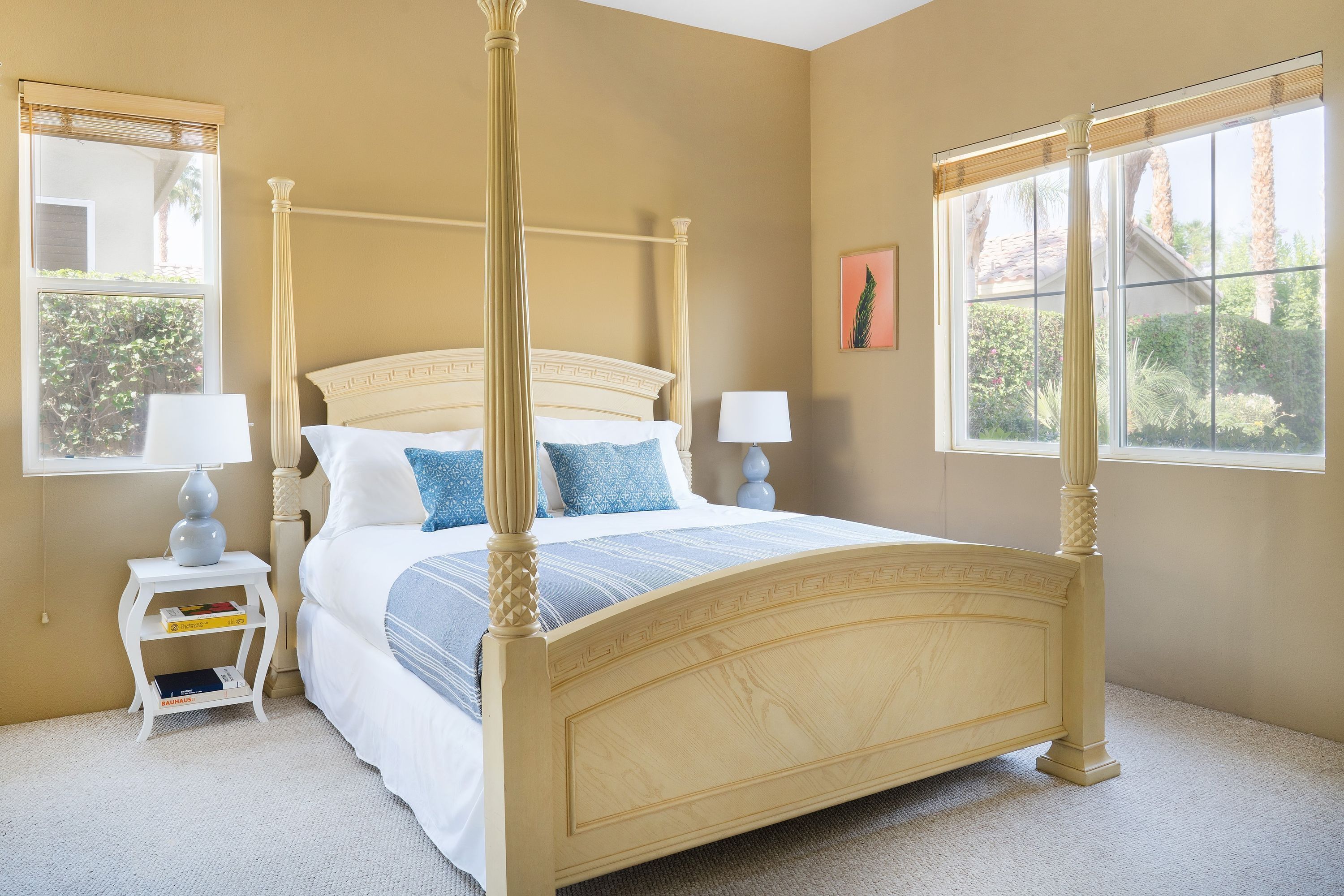 A four-poster bed with a white headboard and footboard, flanked by two bedside tables with lamps, in a room with beige walls and large windows.