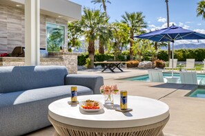A poolside area with a round table, a sofa, and a patio umbrella. - Polo Villa 3 by Avantstay Features Expansive Putting Green Pool Spa Outdoor Firepit 260328 6 Bedrooms (La Quinta)