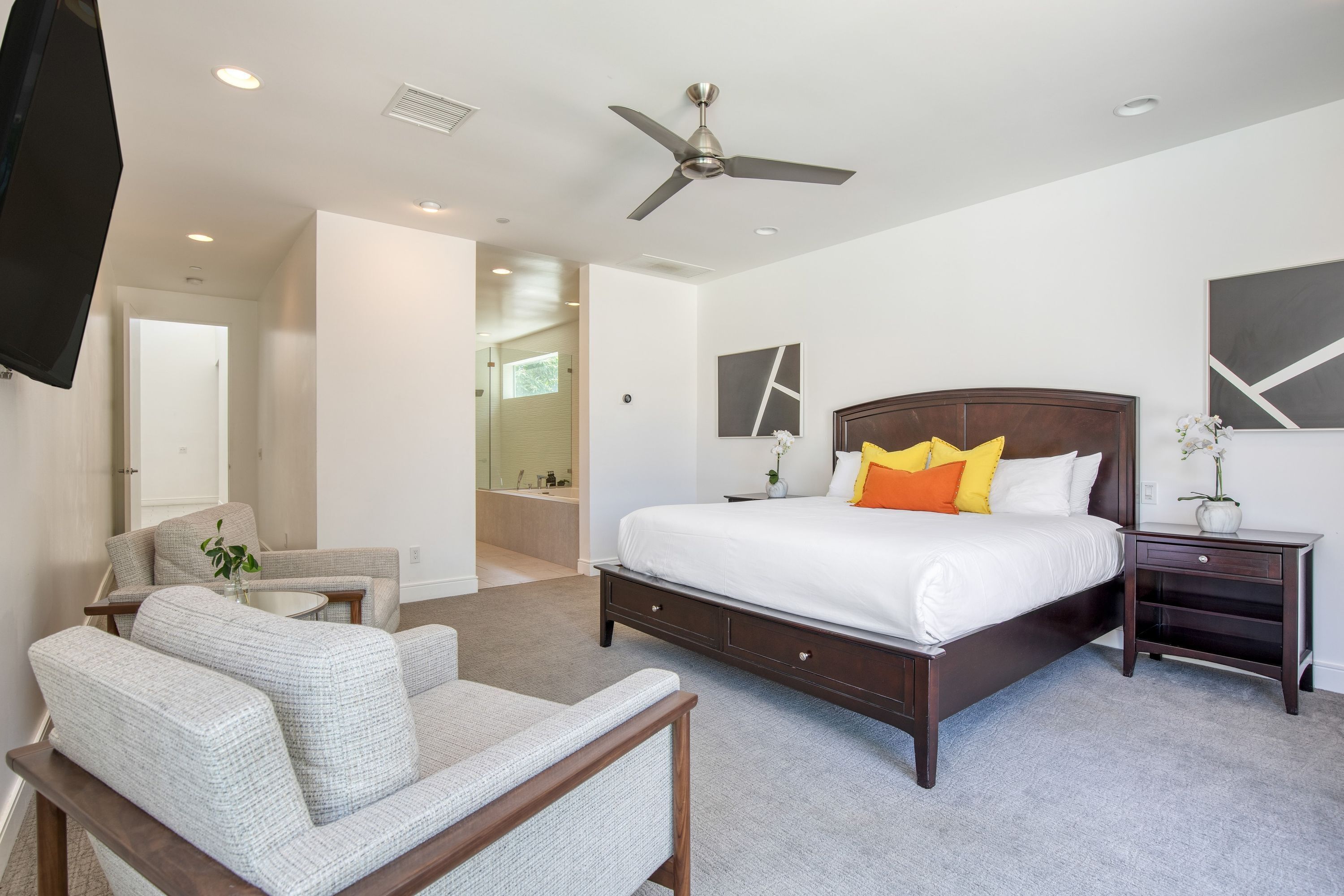 A bedroom with a large bed, a chair, a bedside table, a ceiling fan, and a TV mounted on the wall.