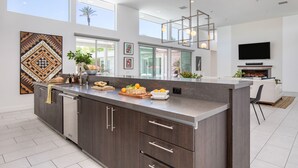 A modern kitchen with a large island, dark cabinetry, and a built-in oven. There are large windows providing natural light and a view of the outdoors.