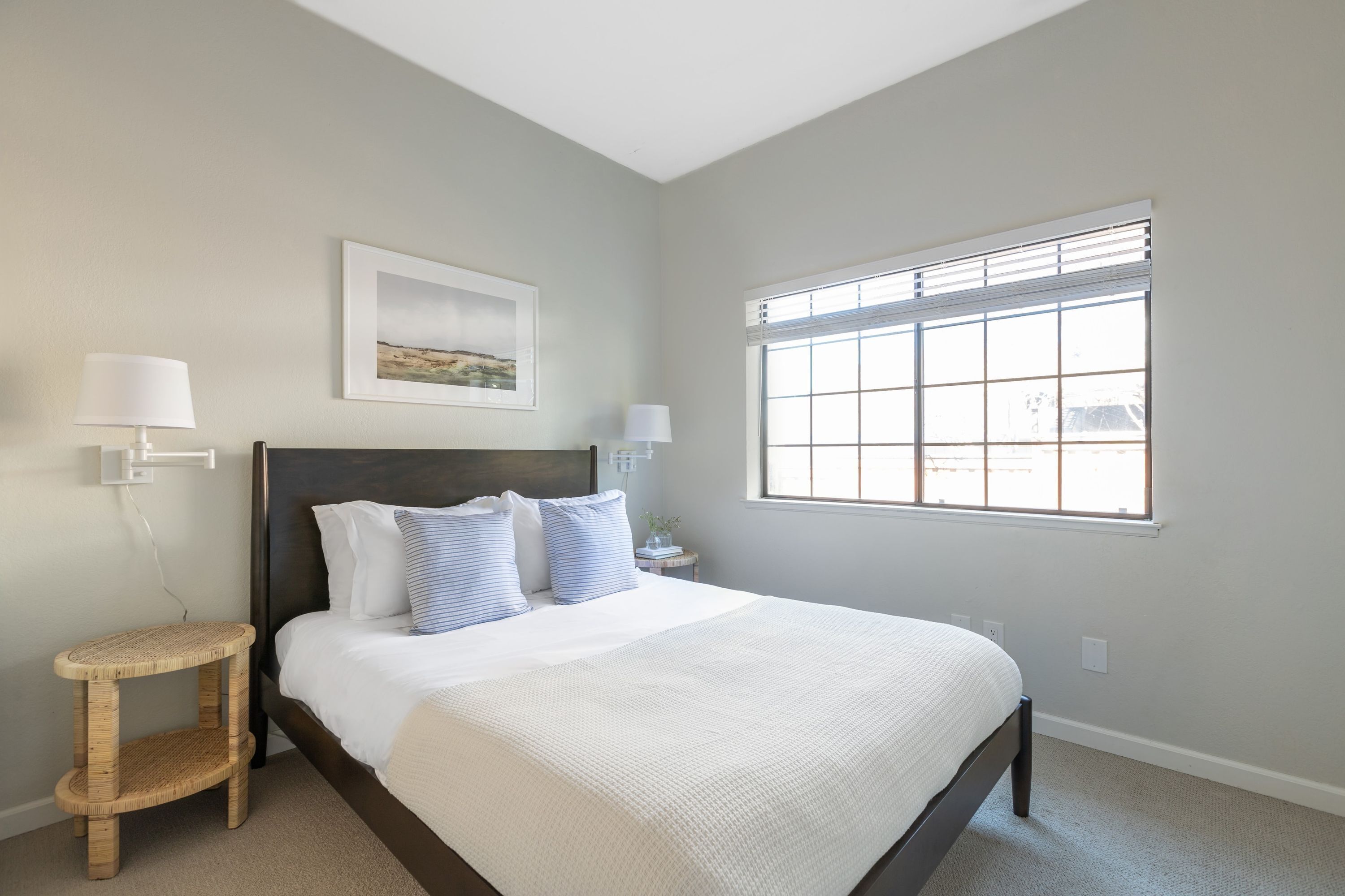 A bedroom with a bed, a window with blinds, a bedside table with a lamp, and a wall-mounted picture.