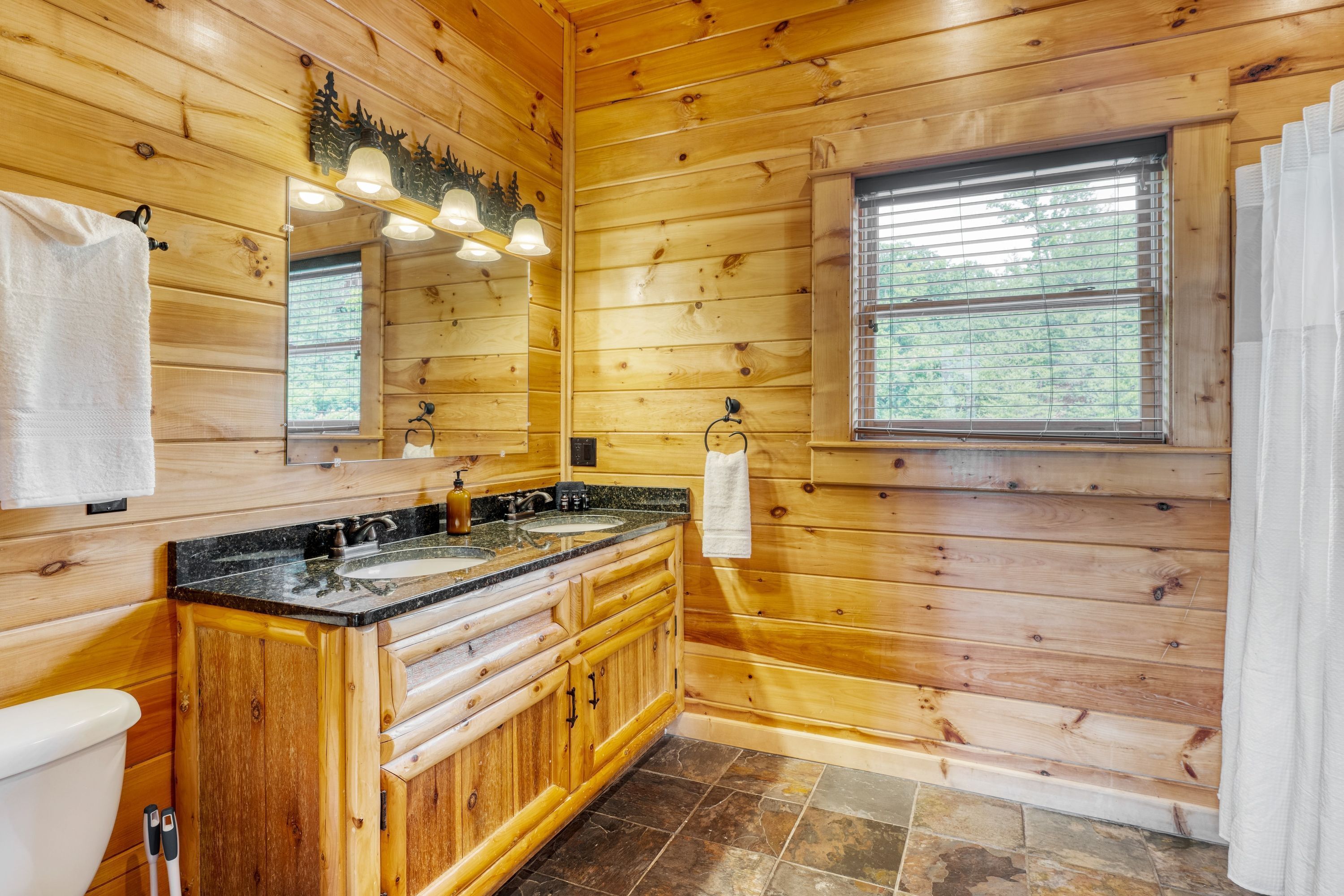 A wooden cabin bathroom with a double sink vanity, a mirror, and a window with blinds.
