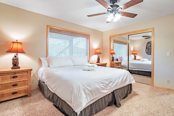 A bedroom with a large bed, two bedside tables with lamps, a ceiling fan, a large window with blinds, and a mirror on the wall.