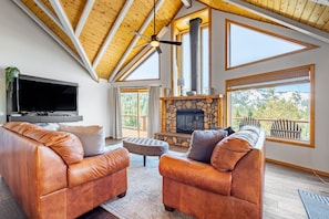 A spacious living room with leather sofas, a stone fireplace, and large windows offering a view of the outdoors. - Endless View Lodge by Avantstay Incredible Views Close To Bear Mountain Ski Resort w Hot Tub (Big Bear Lake)